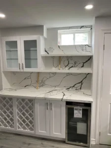 Basement wet bar with white shaker lower cabinets brushed nickel hardware dark veined quartz countertop and full height backsplash diamond wine rack glass front upper cabinet floating shelves and stainless beverage fridge