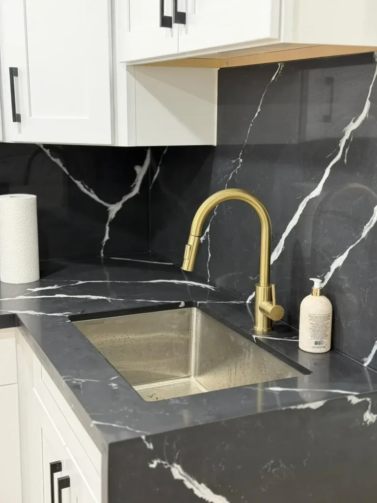 Closeup of black quartz countertop undermount sink with gold faucet and black slab backsplash by MD Kitchen and Bath