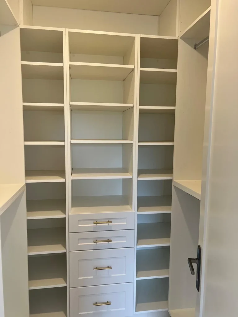 Custom white walk-in closet with floor to ceiling open shelving on both sides center tower with four shaker drawers gold brass bar pull hardware and chrome hanging rod on right side by MD Kitchen and Bath