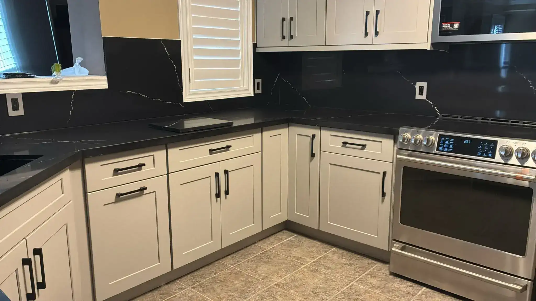 L-shape grey shaker kitchen with black Nero quartz countertop and full height backsplash matte black hardware stainless electric range plantation shutters and beige tile floor by MD Kitchen and Bath
