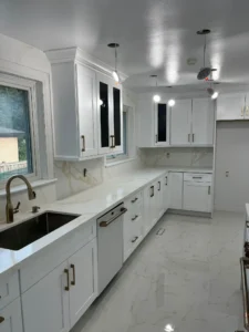 After photo of renovated kitchen showing white shaker upper cabinets with glass door inserts brushed bronze bar pulls black undermount sink brushed bronze gooseneck faucet calacatta gold quartz countertop and backsplash stainless dishwasher and large format white marble porcelain tile floor