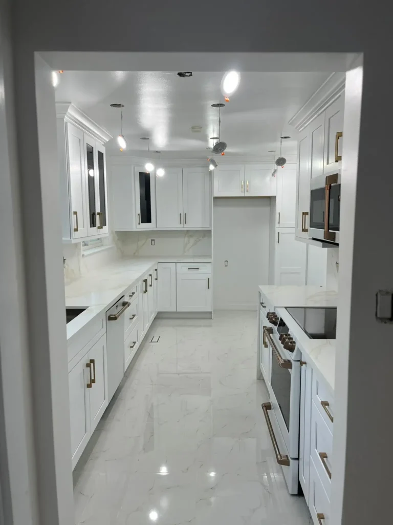 After photo of renovated galley kitchen viewed from entrance doorway showing full corridor layout with white shaker cabinets brushed bronze bar pulls calacatta gold quartz countertops and backsplash Cafe white range and microwave glass door upper cabinets large format white marble porcelain tile floor and recessed pot lights