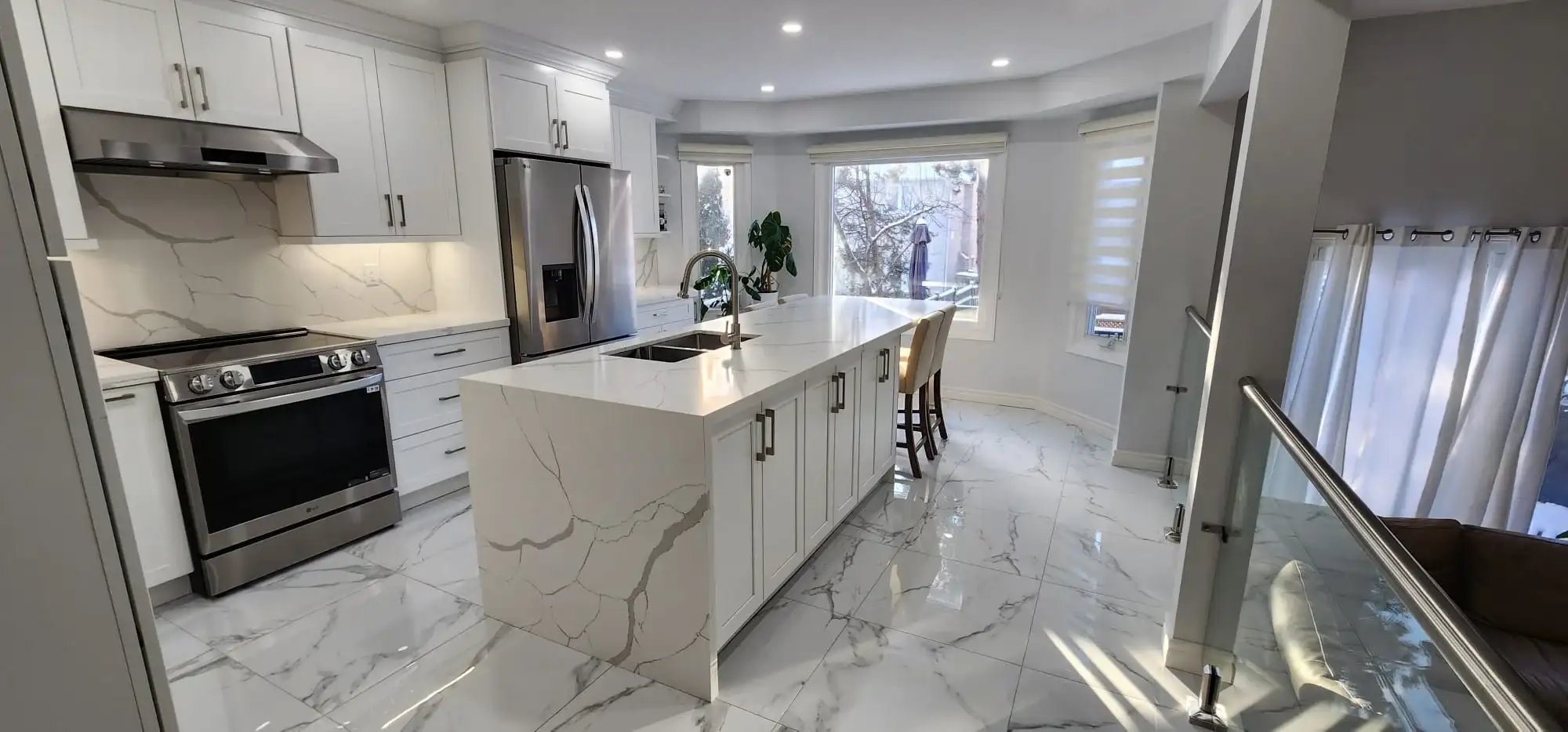 After photo of renovated kitchen taken from staircase angle showing LG stainless gas range white shaker cabinets calacatta quartz island and countertops matching quartz backsplash stainless range hood glass panel railing and marble porcelain tile floor