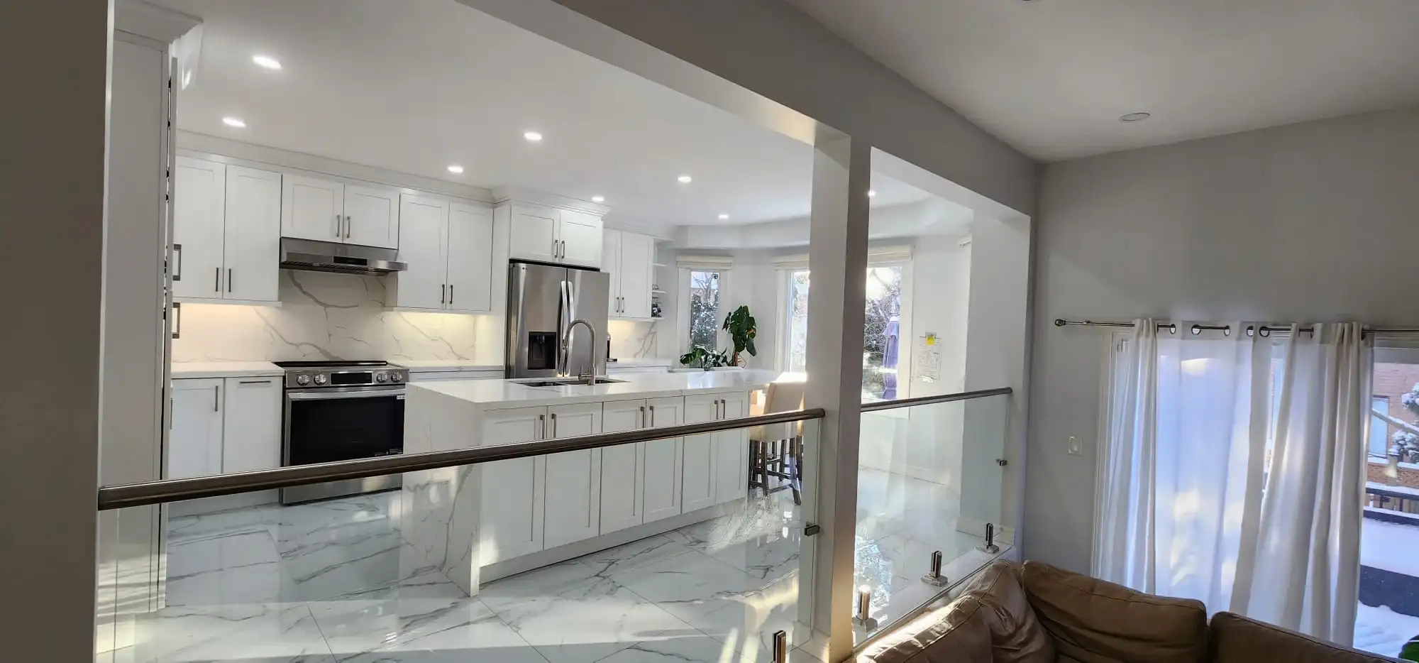 After photo showing renovated white shaker kitchen calacatta quartz island and marble look porcelain tile floor viewed from living room through modern glass panel staircase railing with dark wood handrail
