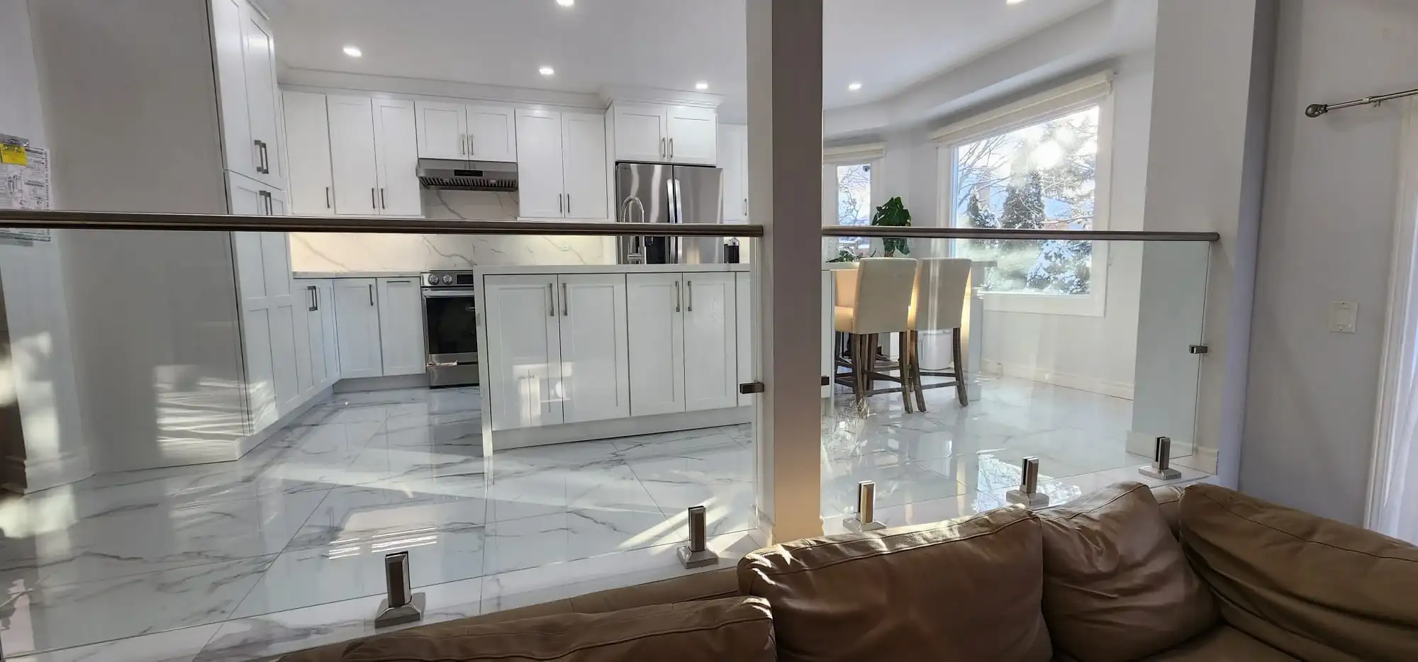 After photo showing completed open concept renovation with modern glass panel staircase railing leather sofa white shaker kitchen calacatta quartz island and marble look porcelain tile floor visible in background