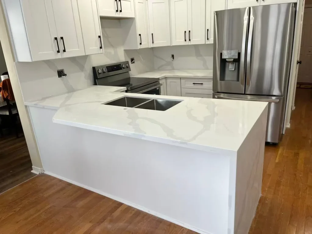 After photo of renovated kitchen showing white shaker upper and lower cabinets matte black bar pulls calacatta quartz peninsula with stainless undermount sink matching quartz backsplash stainless Samsung electric range stainless French door refrigerator with water dispenser and hardwood flooring