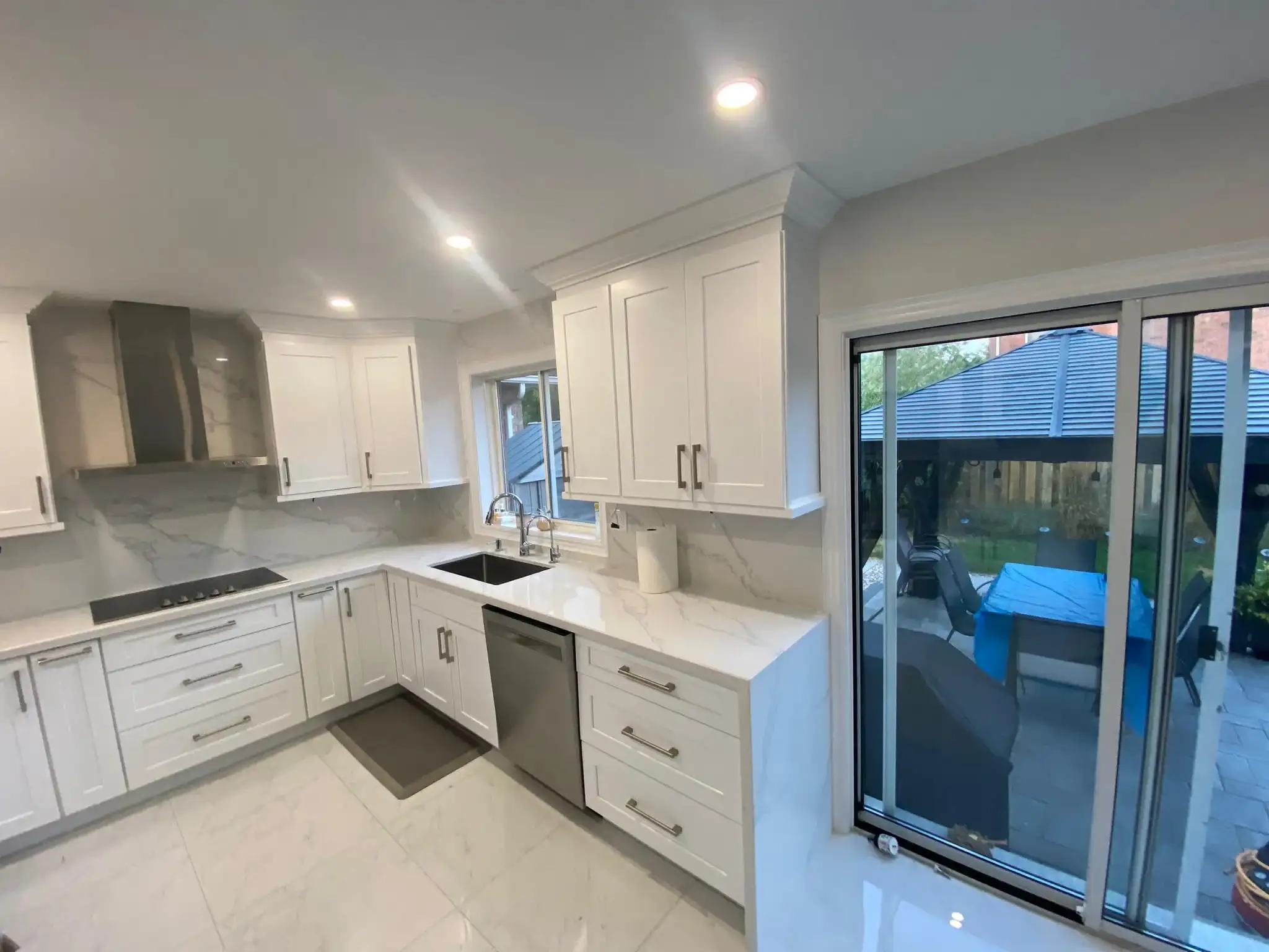 After photo of renovated kitchen showing white shaker upper and lower cabinets calacatta quartz countertop and backsplash stainless wall mount range hood black cooktop stainless dishwasher brushed nickel bar pulls large format porcelain tile floor and sliding glass door to backyard