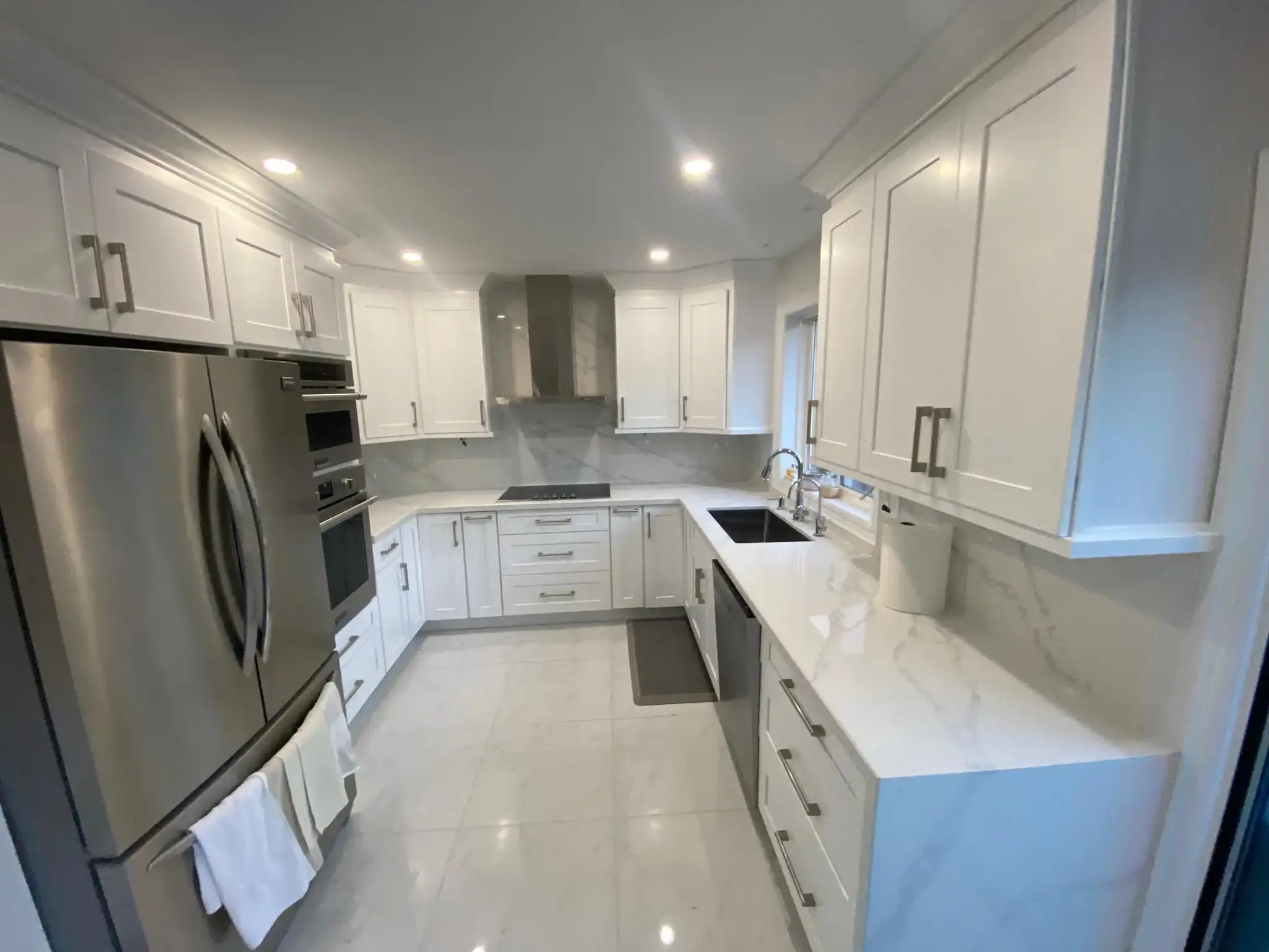 After photo of full U-shape renovated kitchen showing white shaker upper and lower cabinets calacatta quartz countertops and backsplash stainless French door refrigerator stainless double wall oven black cooktop stainless wall mount range hood brushed nickel bar pulls and large format porcelain tile floor