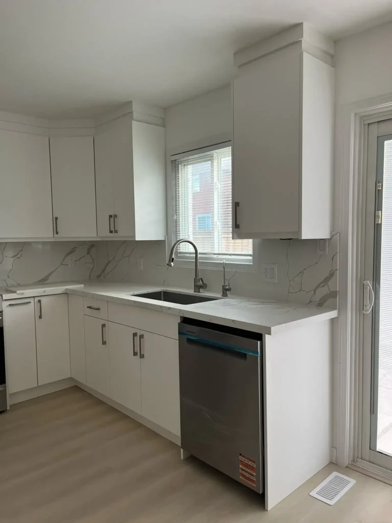 After photo of renovated kitchen sink wall showing white flat panel slab upper and lower cabinets black undermount sink brushed nickel gooseneck faucet stainless dishwasher calacatta quartz countertop and backsplash sliding patio door and light gray LVP flooring