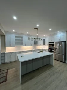Wide angle after photo of completed kitchen renovation showing full two tone layout with light blue gray shaker perimeter cabinets dark charcoal island calacatta quartz countertops and backsplash brass hardware recessed pot lights brass pendant light stainless appliances and light oak LVP flooring