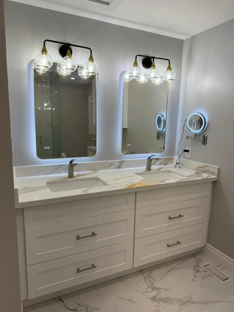 Master bathroom double vanity with white shaker cabinets brushed nickel hardware Calacatta gold quartz countertop dual undermount sinks two LED backlit mirrors gold and black wall sconces and large format Calacatta marble tile floor by MD Kitchen and Bath