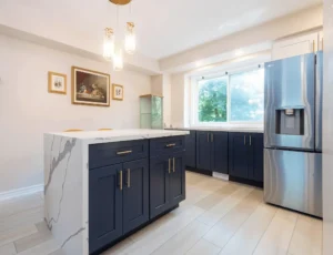 Navy blue shaker lower cabinets and island with brushed gold bar pulls calacatta quartz waterfall island top white upper shelving unit LG stainless French door refrigerator with water dispenser crystal drop pendant light light wood look porcelain tile floor and framed artwork on wall