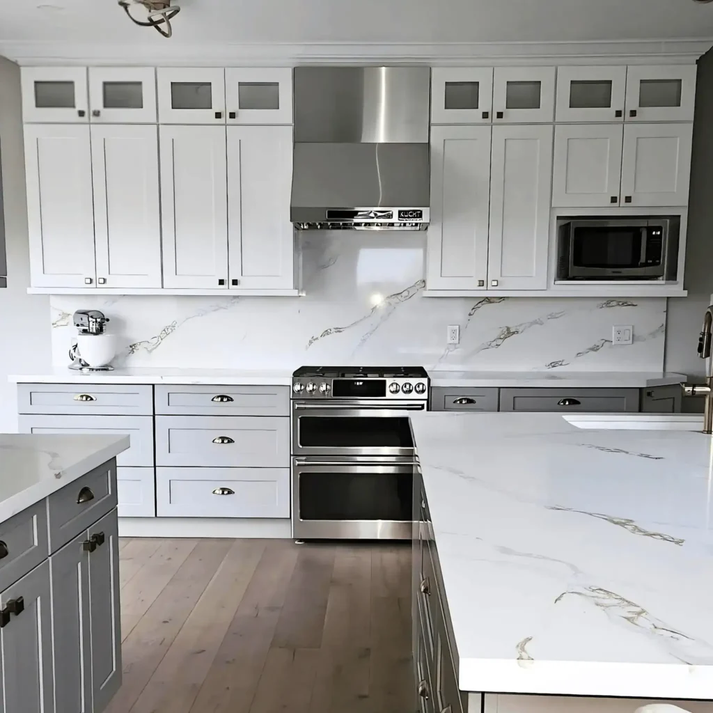 Two tone kitchen with white shaker upper cabinets featuring glass inserts and gray shaker lower cabinets with antique brass cup pulls stainless double oven range Kucht stainless chimney range hood calacatta gold quartz countertop and backsplash built-in microwave and light oak hardwood flooring