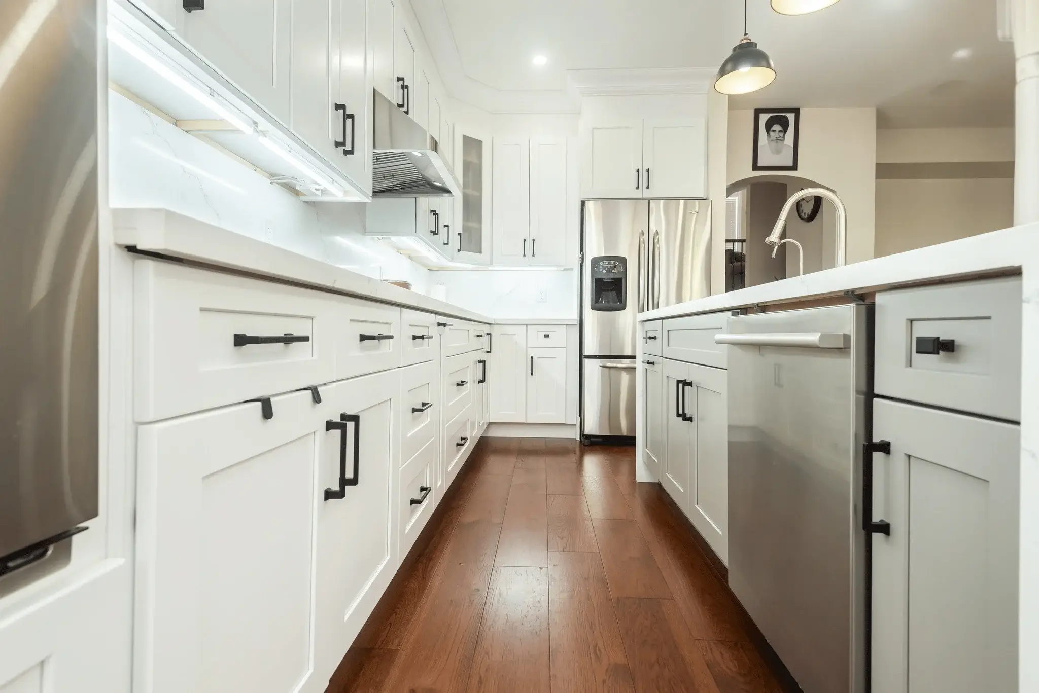 Wide galley view of white shaker upper and lower cabinets with matte black bar pulls calacatta quartz countertop and backsplash under cabinet lighting stainless range hood stainless French door refrigerator with water dispenser stainless dishwasher and dark hardwood flooring