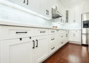 Closeup of white shaker lower cabinets and drawers with matte black bar pulls calacatta quartz countertop and backsplash under cabinet LED lighting stainless range hood and dark hardwood flooring