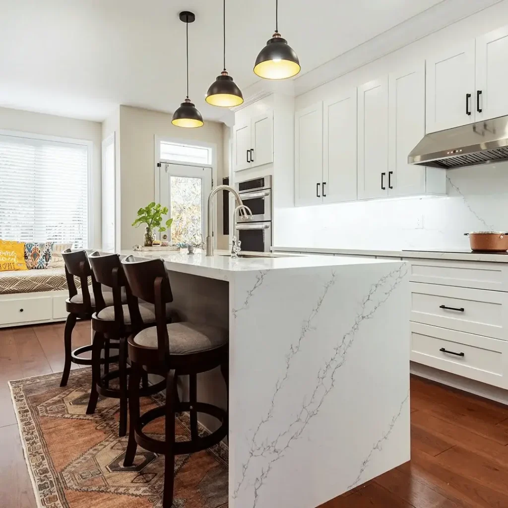 White shaker upper and lower cabinets matte black bar pulls calacatta quartz waterfall island with stainless sink dark walnut swivel bar stools three dark dome pendant lights stainless double wall oven stainless range hood and dark hardwood flooring