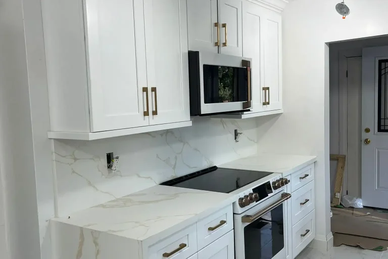 After photo of renovated kitchen featuring white shaker cabinets with crown moulding Cafe white electric range with brushed bronze handles over-range microwave brushed bronze bar pulls calacatta gold quartz countertop and full height backsplash and large format white marble porcelain tile floor