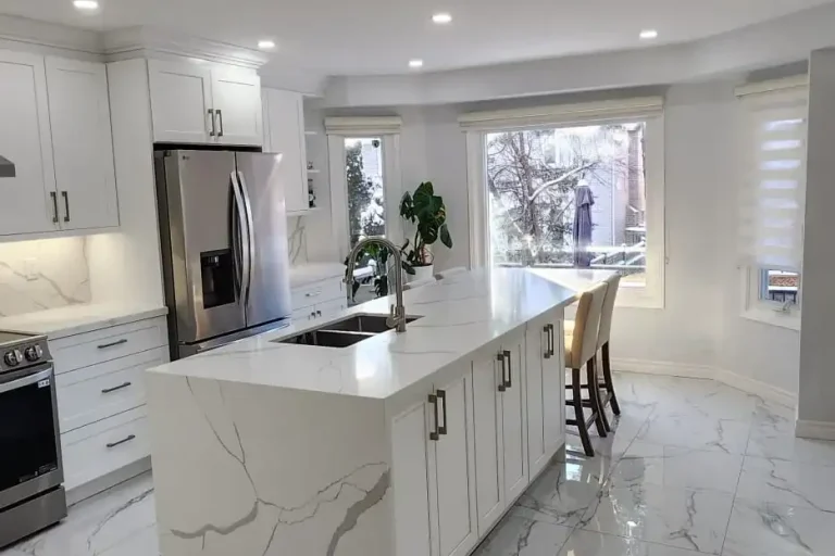 Wide after photo of renovated kitchen showing white shaker cabinets calacatta quartz island with waterfall sides black undermount sink stainless LG refrigerator large format marble look porcelain tile floor and large windows
