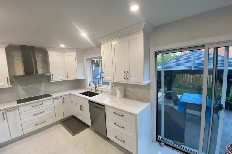 After photo of renovated kitchen showing white shaker upper and lower cabinets calacatta quartz countertop and backsplash stainless wall mount range hood black cooktop stainless dishwasher brushed nickel bar pulls large format porcelain tile floor and sliding glass door to backyard