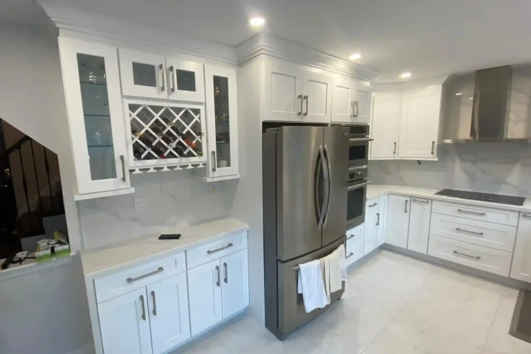 After photo of renovated kitchen showing custom white shaker upper cabinets with glass doors built-in diamond wine rack stainless French door refrigerator stainless double wall oven calacatta quartz countertop and backsplash brushed nickel bar pulls and large format porcelain tile floor