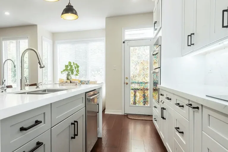 White shaker upper and lower cabinets matte black bar pulls calacatta quartz island with stainless undermount sink two dark dome pendant lights stainless dishwasher dark hardwood flooring and back door entry with natural light
