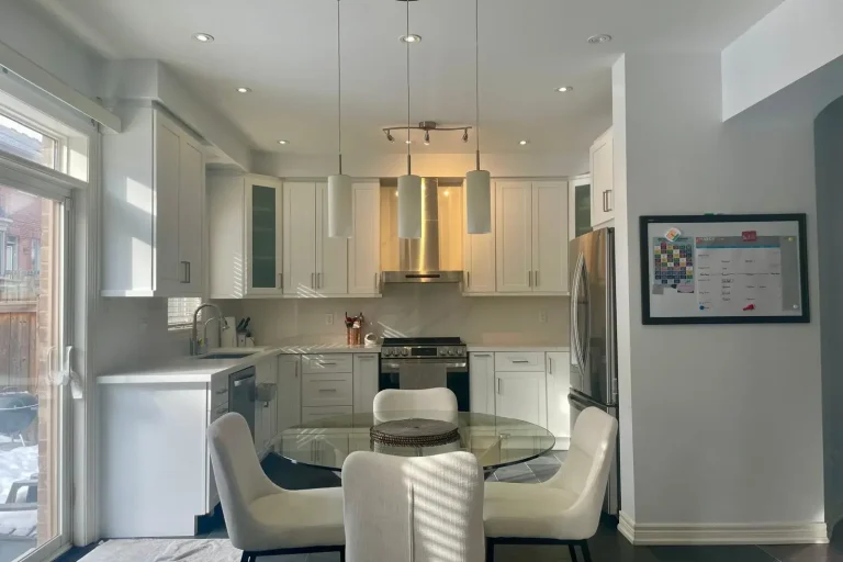 White shaker kitchen with glass front uppers stainless chimney hood cylinder pendant lights round glass dining table white upholstered chairs and dark porcelain tile floor by MD Kitchen and Bath