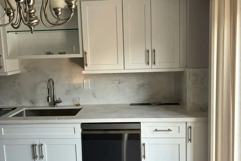 White shaker kitchen sink wall with brushed nickel hardware stainless undermount sink brushed nickel faucet cloud grey quartz backsplash stainless dishwasher silver chandelier dark charcoal island in foreground and dark hardwood floor by MD Kitchen and Bath