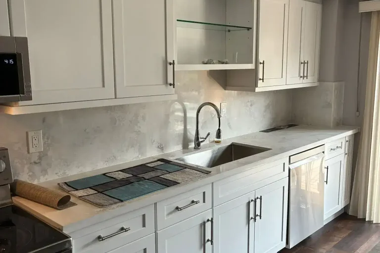 White shaker kitchen sink wall with brushed nickel bar pull hardware stainless undermount sink brushed nickel faucet cloud grey quartz backsplash white quartz countertop stainless dishwasher glass shelf display upper and dark hardwood floor by MD Kitchen and Bath