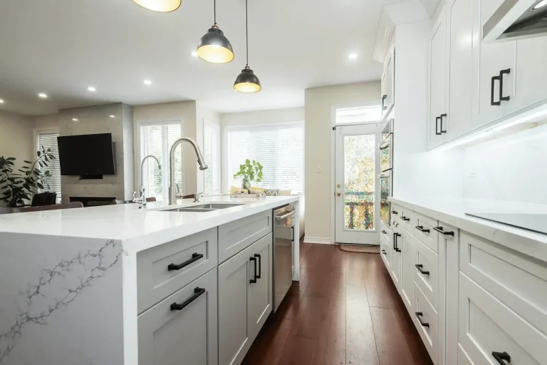 White shaker kitchen with two tone gray island base calacatta quartz waterfall countertop stainless undermount sink matte black bar pulls dark industrial pendant lights under cabinet lighting and dark hardwood flooring
