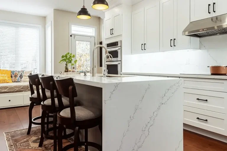 White shaker upper and lower cabinets matte black bar pulls calacatta quartz waterfall island with stainless sink dark walnut swivel bar stools three dark dome pendant lights stainless double wall oven stainless range hood and dark hardwood flooring